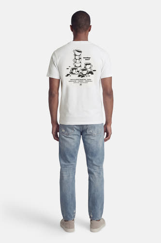 Wit T-shirt van KAOTIKO, met zwarte koffiekopjes print op de achterkant, gecombineerd met blauwe jeans en suède schoenen.
