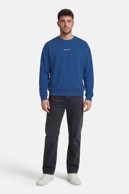 Blauwe sweater van KAOTIKO, met een witte tekst op de borst, gecombineerd met een donkere jeans en witte sneakers.