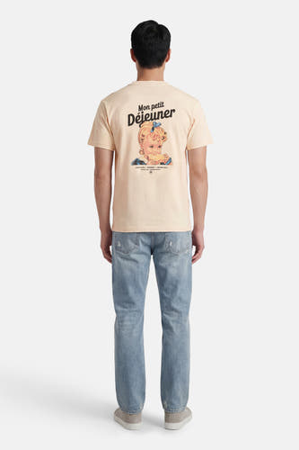 T-shirt écru de KAOTIKO, avec un imprimé d'une fille portant le texte "Mon petit Déjeuner" dans le dos, porté avec un jean et des baskets.
