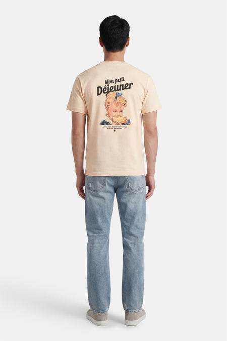 Ecru T-shirt van KAOTIKO, met opdruk van een meisje met de tekst "Mon petit Déjeuner" op de rug, gedragen met jeans en sneakers.