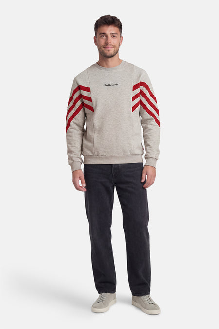 Grijze sweater van KAOTIKO, met ronde hals en rode strepen op de mouwen, gecombineerd met een donkere jeans en grijze schoenen.
