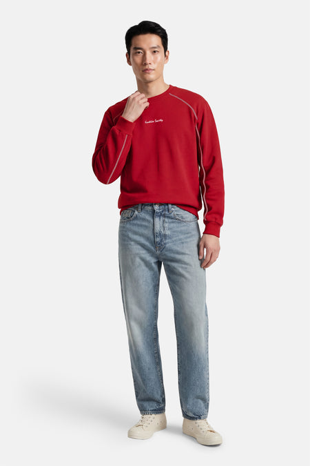 Rode sweater met ronde hals van Kaotiko, afgewerkt met witte stiksels, gecombineerd met een blauwe jeans en crèmekleurige sneakers.
