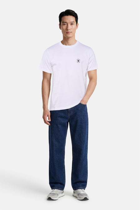 T-shirt blanc KAOTIKO avec petit logo sur la poitrine, porté par un mannequin avec un jean bleu foncé et des baskets grises.