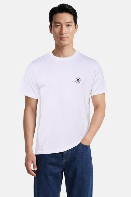 T-shirt blanc à manches courtes de KAOTIKO avec un petit logo rond noir avec une empreinte de patte et 'KAOTIKO SOCIETY' sur la poitrine, porté avec un jean bleu foncé.