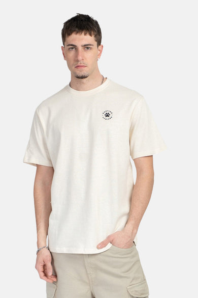 T-shirt écru de KAOTIKO, avec un petit logo sur la poitrine, porté avec un pantalon cargo kaki.