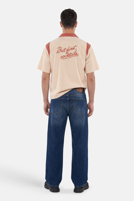 Multicolor overhemd met korte mouwen van KAOTIKO, beige met terracotta details en 'But first, cocktails' op de rug, gedragen door een man met een donkere jeans.