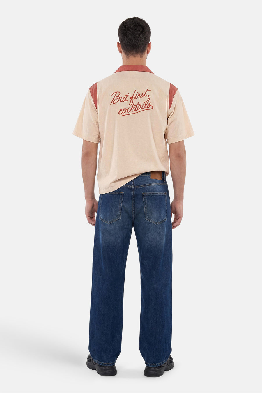 Multicolor overhemd met korte mouwen van KAOTIKO, beige met terracotta details en 'But first, cocktails' op de rug, gedragen door een man met een donkere jeans.