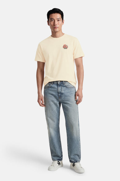 T-shirt écru de KAOTIKO, à manches courtes et avec un petit imprimé sur la poitrine, combiné avec un jean bleu et des baskets blanches.