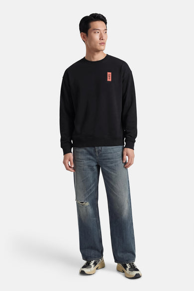 Pull noir de KAOTIKO, avec un patch rectangulaire vertical rouge et porté avec un jean bleu.