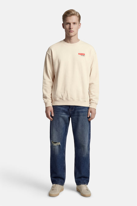 Ecru sweater met ronde hals van KAOTIKO, met rode tekstprint op de borst, gedragen met blauwe jeans met gat.