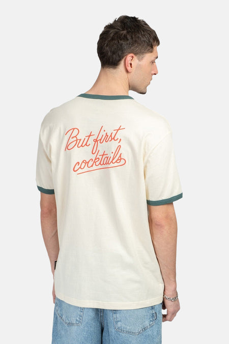 T-shirt écru à manches courtes de KAOTIKO, avec finition verte et le texte "But first, cocktails" dans le dos.