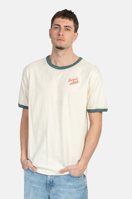 T-shirt écru de KAOTIKO avec bords verts et inscription 'But first cocktails', porté avec un jean.