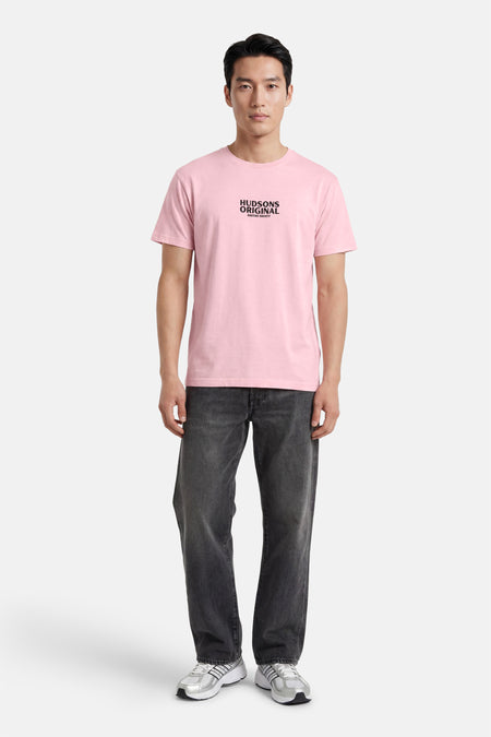 T-shirt rose de KAOTIKO avec imprimé texte noir sur la poitrine, porté par un homme avec un jean foncé et des baskets argentées.