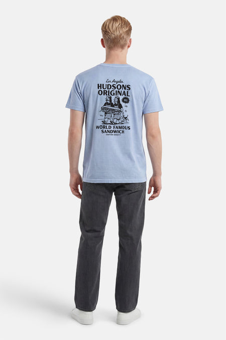 Lichtblauw T-shirt van KAOTIKO met een grote zwarte grafische print op de rug van twee figuren die een broodje dragen, gedragen door een man met donkergrijze broek en witte sneakers.