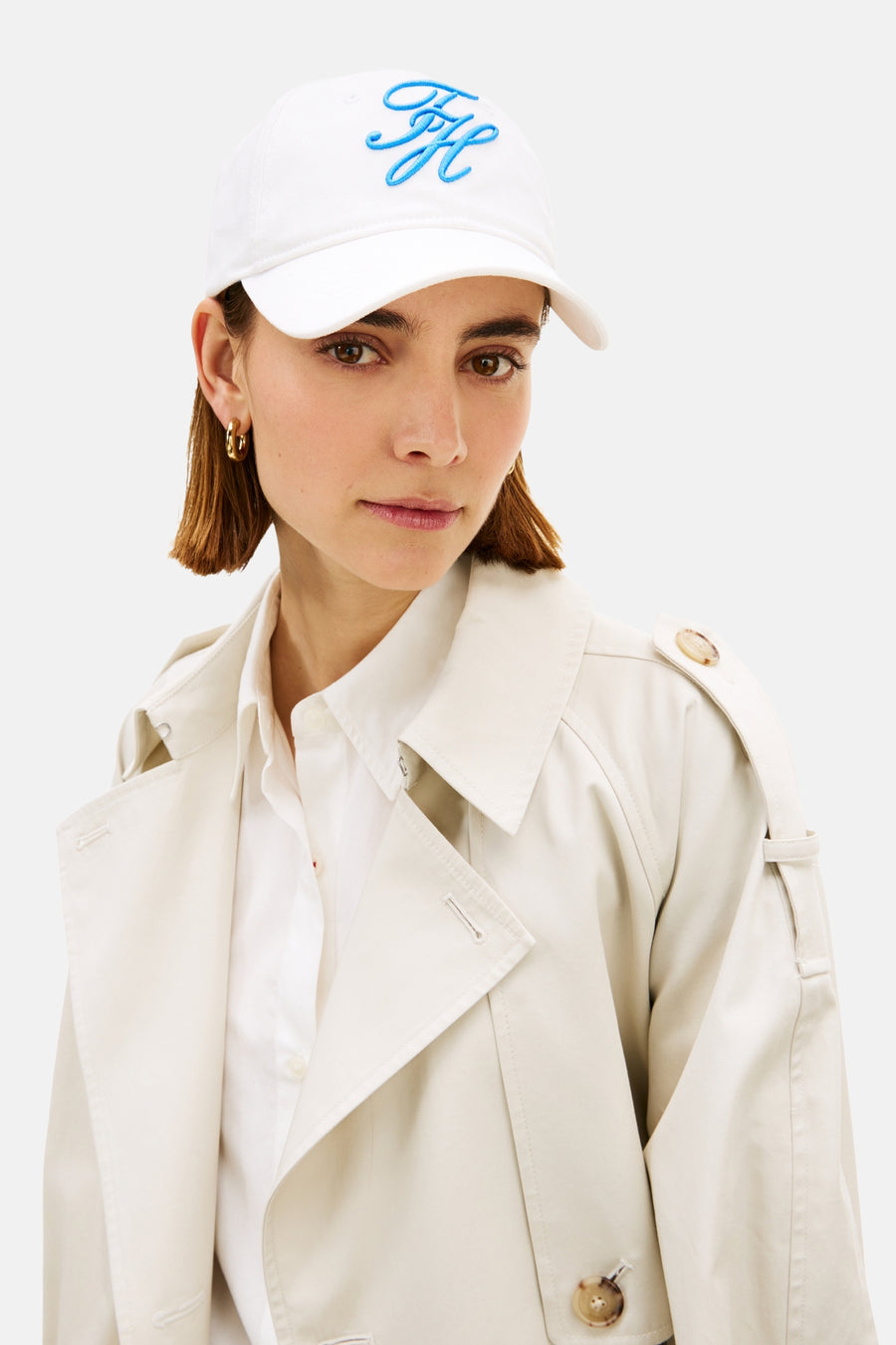 Witte pet van TOMMY JEANS, met blauw geborduurd logo op de voorkant, gedragen met een trenchcoat en een wit overhemd.