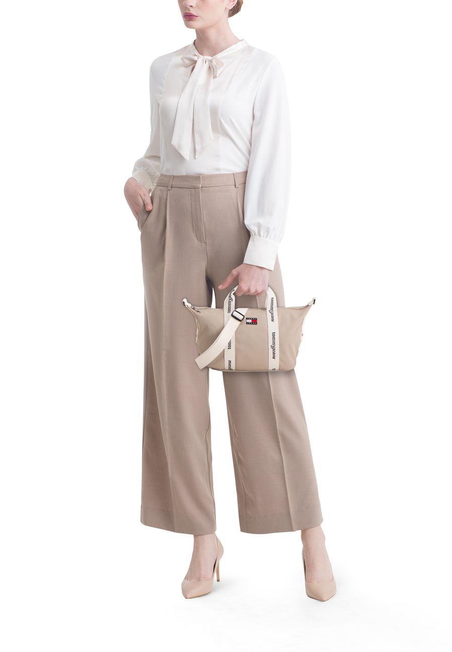 Sac à bandoulière beige de TOMMY JEANS, avec sangles et logo blancs, porté avec une blouse blanche et un pantalon beige.