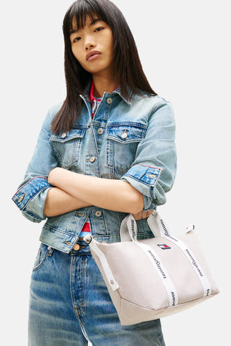 Sac à bandoulière beige de Tommy Jeans, avec des sangles blanches et un logo, porté avec une veste en jean et un jean.