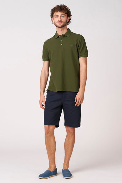 Blauwe bermuda van Bruce & Butler, met een effen dessin en gecombineerd met een olijfgroen poloshirt en blauwe espadrilles.
