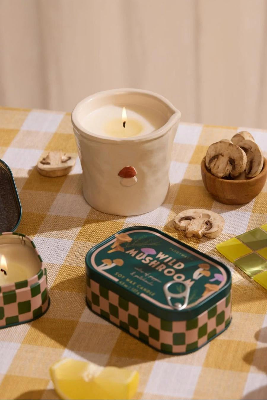 Groene kaars in metalen blik met schaakbordpatroon van PADDY WAX, op gele geruite tafelkleed, naast houten kom met paddenstoelen