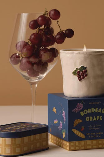 Blauwe kaars in witte pot met druivenmotief van PADDY WAX, brandend, naast wijnglas met druiven en verpakking