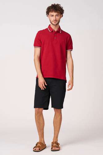 Zwarte short van Bruce & Butler, gecombineerd met een rood poloshirt met witte details en suède sandalen.
