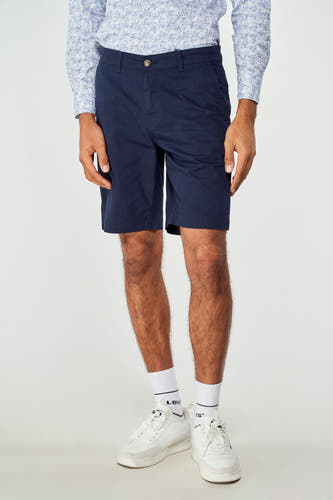 Blauwe bermuda van Bruce & Butler, gedragen met een overhemd met print en witte sneakers met sokken.
