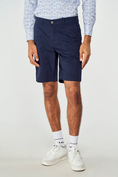 Blauwe bermuda van Bruce & Butler, gedragen met een overhemd met print en witte sneakers met sokken.
