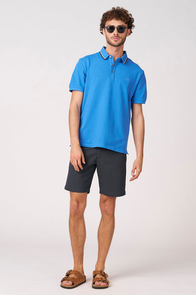 Blauwe short van Bruce & Butler, gedragen met een blauw poloshirt en bruine sandalen.
