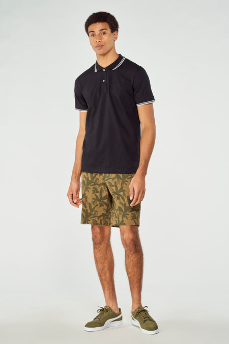 Shorts verts à imprimé palmier de Bruce & Butler, associés à un polo noir et des baskets vertes.