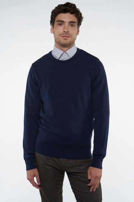 Blauwe trui met ronde hals van Bruce & Butler, gedragen over een overhemd met print en een bruine broek.
