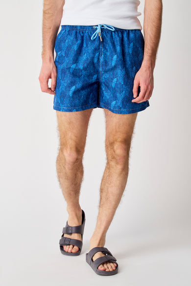 Blauwe zwemshort van Bruce & Butler, met een bladerpatroon en gedragen met grijze sandalen.