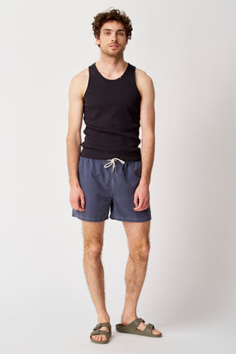 Blauwe zwembroek van Bruce & Butler, gedragen met een zwarte tanktop en groene sandalen.