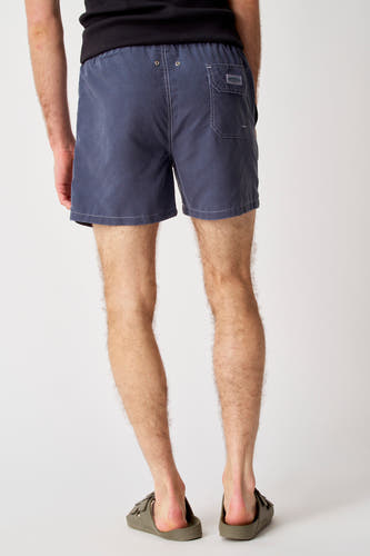 Blauwe zwembroek van Bruce & Butler, gezien van achteren met achterzak en gedragen met kaki sandalen.