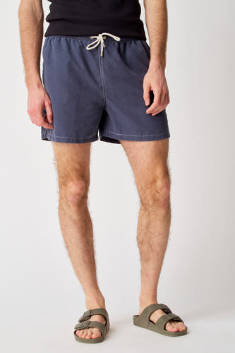 Blauwe zwembroek van Bruce & Butler, met een trekkoord en witte stiksels, gedragen met groene sandalen.
