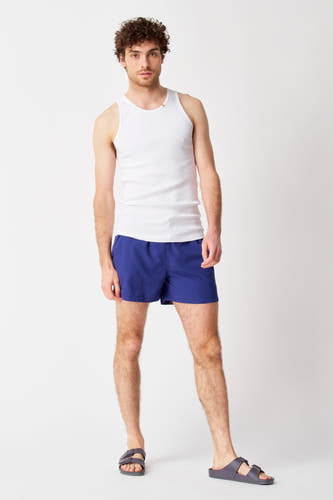 Blauwe zwembroek van Bruce & Butler, gedragen met een wit hemd en grijze sandalen.