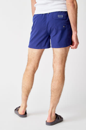 Blauwe zwembroek van Bruce & Butler, gezien van achteren, met achterzak en gedragen met sandalen.