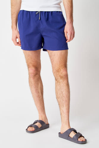 Blauwe zwembroek van Bruce & Butler, gedragen met grijze sandalen en een wit hemd.