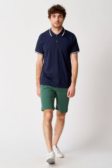 Groene short van Bruce & Butler, gedragen met een donkerblauw poloshirt met witte details en witte sneakers.
