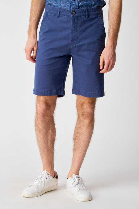 Blauwe short van Bruce & Butler, gedragen met een overhemd met print en witte sneakers.
