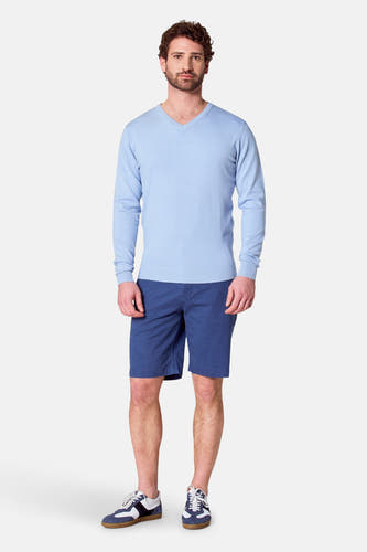 Lichtblauwe trui met V-hals van Bruce & Butler, gedragen met blauwe shorts en sneakers.
