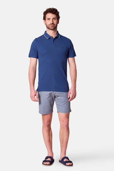 Blauwe polo met korte mouwen van Bruce & Butler, gedragen met grijze shorts en blauwe sandalen.
