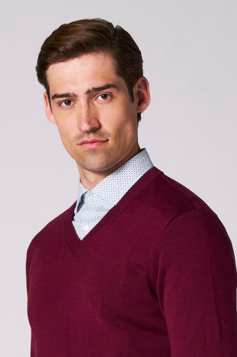 T-shirt V-hals Bordeaux de Bruce & Butler, avec un chemisier bleu clair en dessous, sur une photo en style portrait.