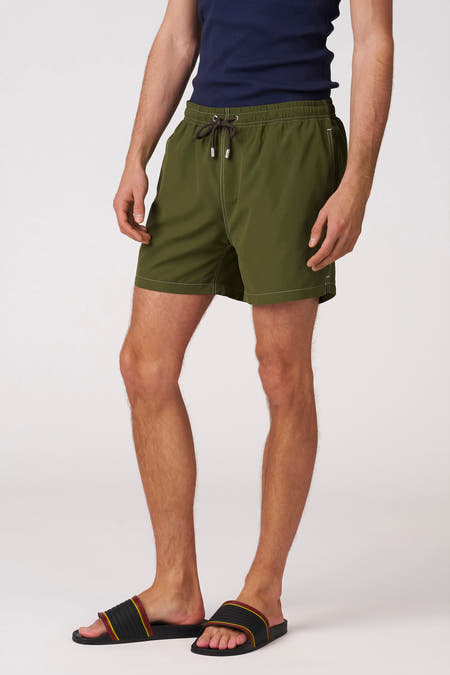 Groene zwemshort van Bruce & Butler met witte stiksels en trekkoord, gedragen door een man met donkerblauwe tanktop en slippers.