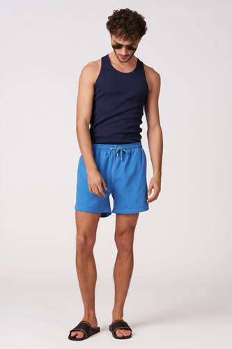 Short de bain bleu de Bruce & Butler avec cordon de serrage et coutures blanches, porté par un homme avec un débardeur bleu marine, des lunettes de soleil et des tongs noires.