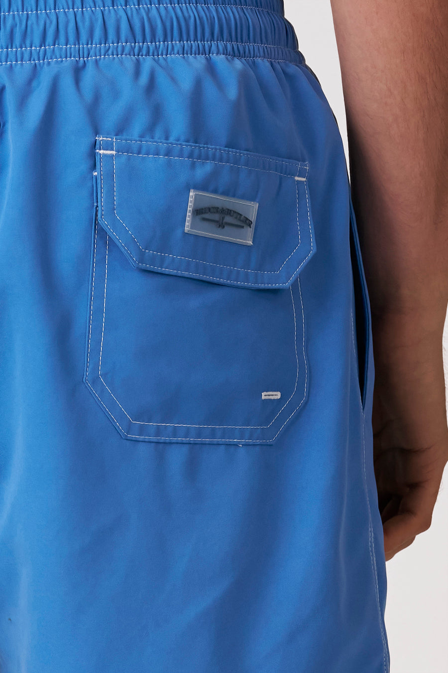Arrière d'un short de bain bleu Bruce & Butler avec taille élastique, coutures contrastées blanches, une poche arrière à rabat et une étiquette transparente.