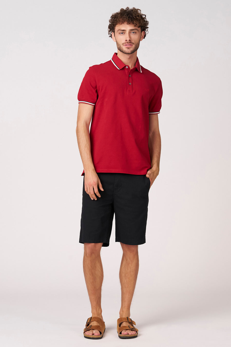 Polo rouge à manches courtes de Bruce & Butler, fini avec des détails blancs, combiné avec un short noir et des sandales.