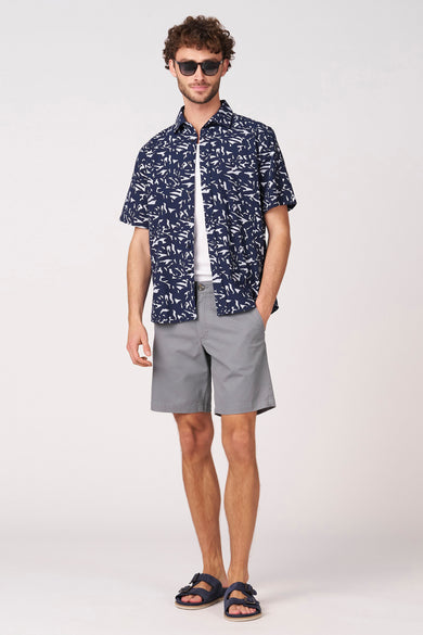 Chemise bleue à manches courtes de Bruce & Butler, avec motifs blancs, combinée à un t-shirt blanc et un short gris.