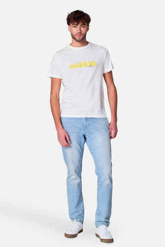 Man staat, witte t-shirt met gele print, lichtblauwe jeans, witte sneakers.