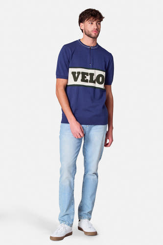 Blauw poloshirt met korte mouwen van Cyclo Club Marcel, met 'VELO' tekst op de borst, gecombineerd met jeans en witte sneakers.
