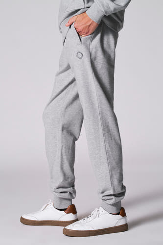 Grijze joggingbroek van Cyclo Club Marcel, met een hand in de zak en witte sneakers met bruine zolen.